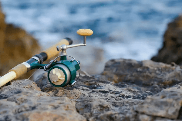 A fishing rod with a green reel resting on rocky ground near the water.