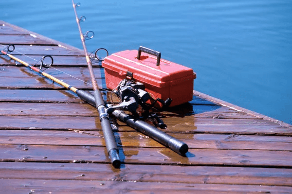 Two fishing rods and a red tackle box sit on a wet wooden dock by calm blue water.