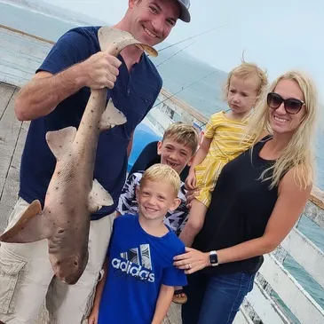 A smiling family poses on a pier; one person holds a large fish, and the ocean is in the background.