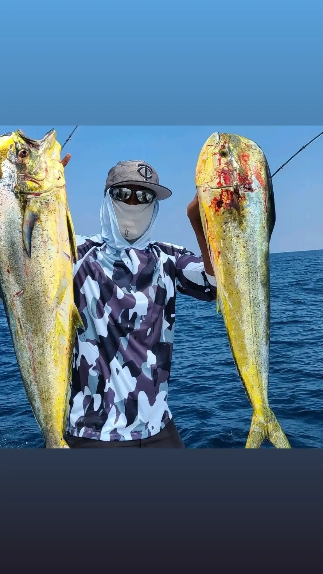 Person in camo shirt and face covering holds two large yellow fish on a boat in the ocean under blue sky.