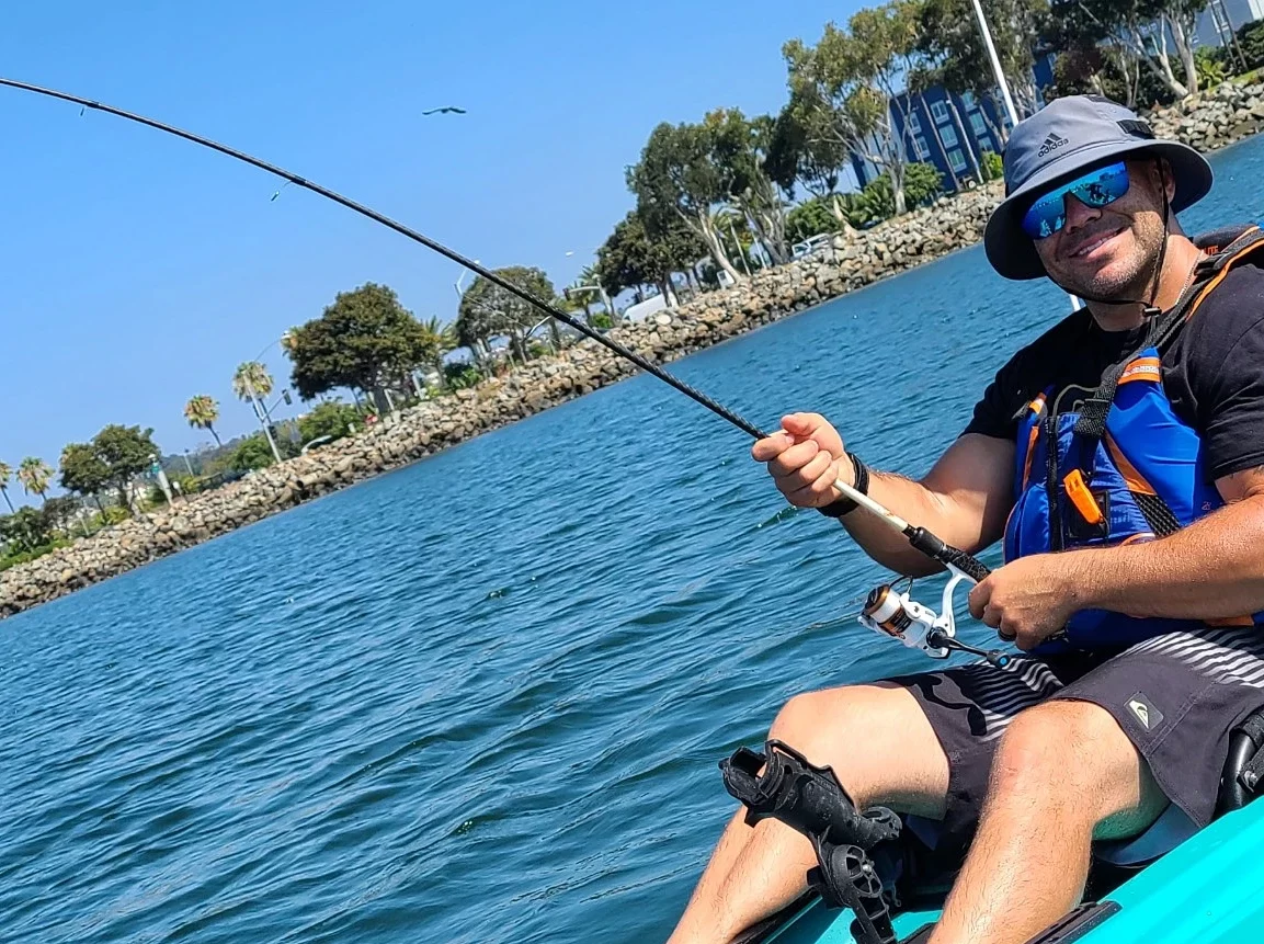 Man fishing on a kayak in calm water, wearing a hat, sunglasses, and life vest, with trees in the background.