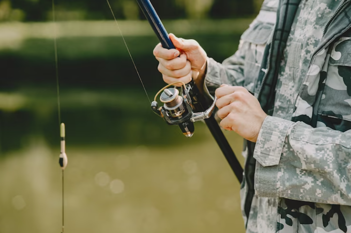 sportfishing Person in camouflage jacket holding a fishing rod, preparing to fish near a body of water.