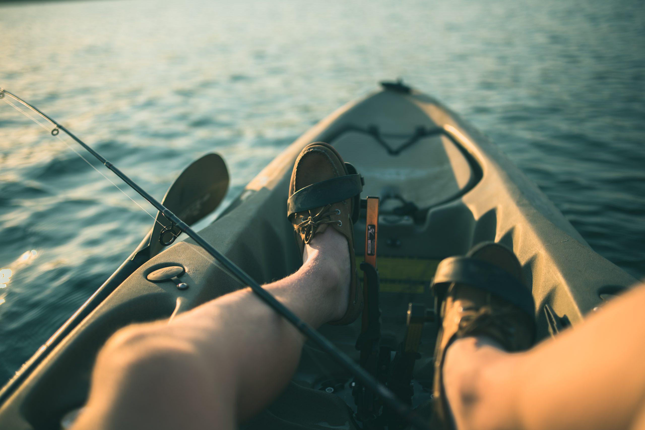 Kayak Fishing Person relaxing in a kayak on calm water, feet up, with a fishing rod resting nearby.