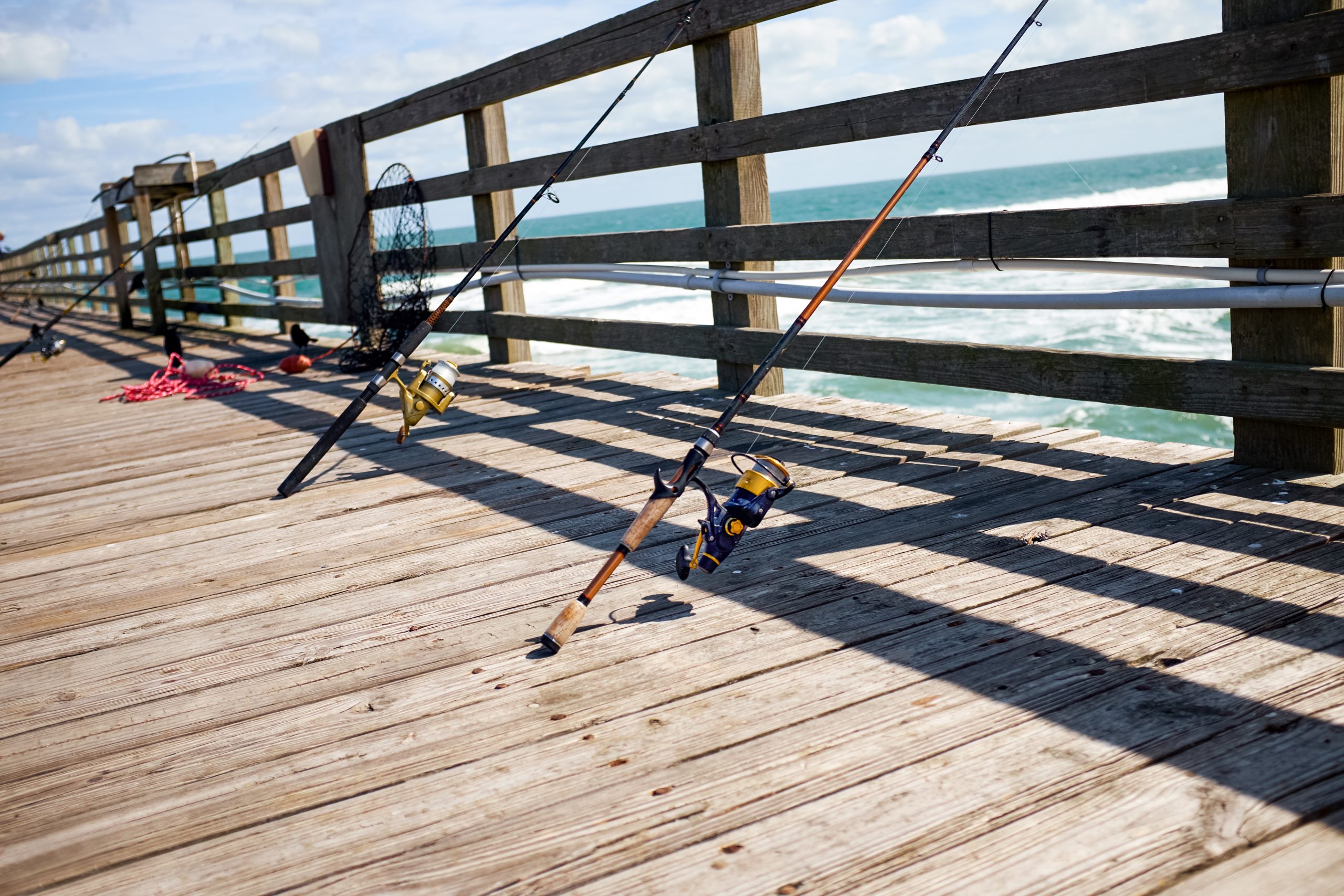 Two fishing poles rest against a wooden pier railing, facing the ocean on a sunny day.