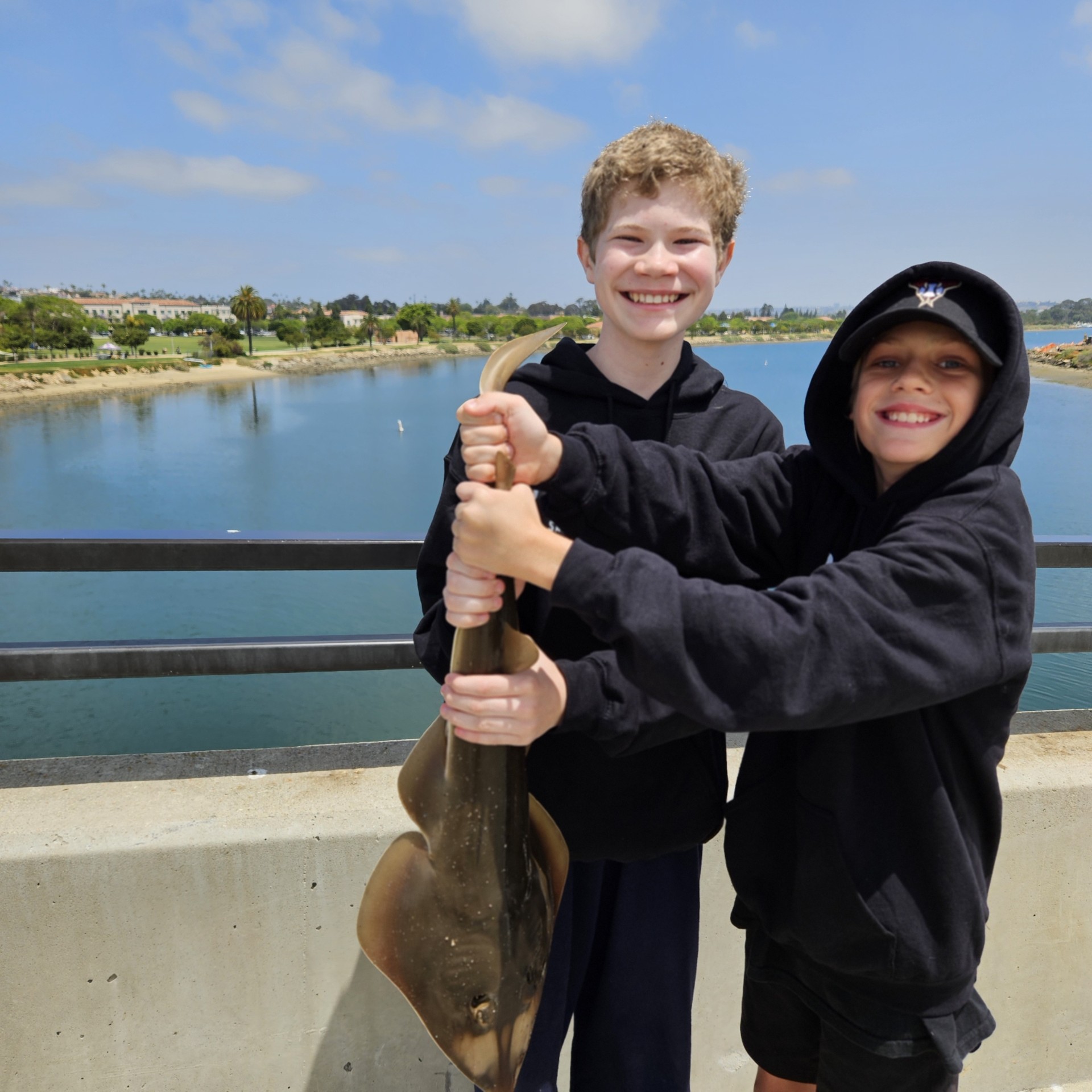 Local Fishing Guide Two smiling kids holding a stingray on a bridge over water, with blue sky and houses in the background.
