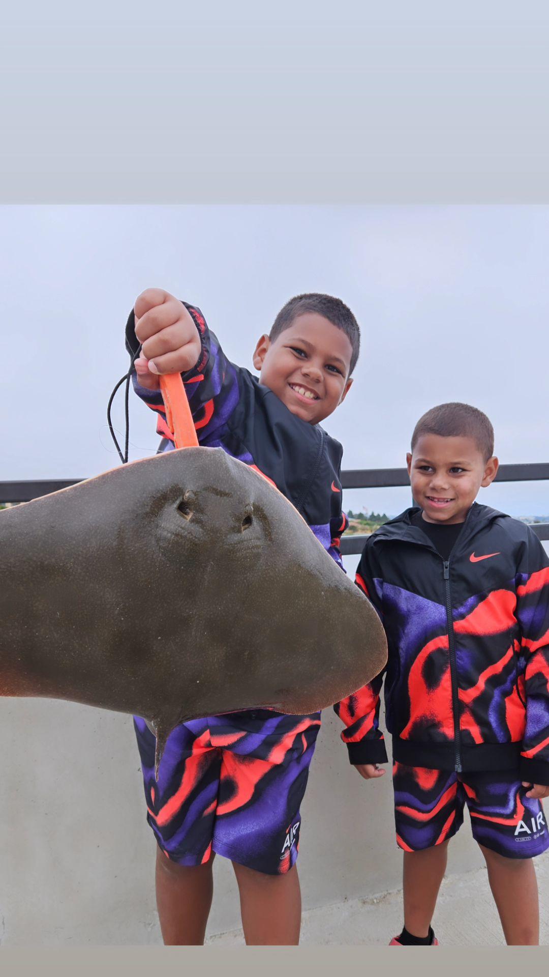 Two boys in matching outfits smile as one holds up a stingray near the water on a cloudy day.