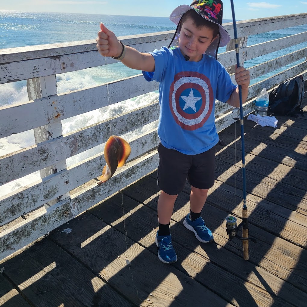 2022-11-25 Boy in a Captain America shirt holding a fishing rod and a small fish on a pier by the ocean.