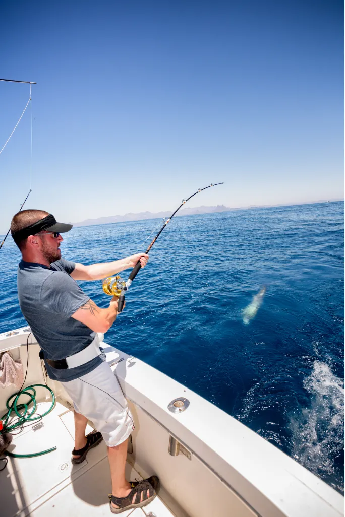 deep-sea-fishing-2 Person fishing from a boat under a clear blue sky, reeling in a catch from the ocean.
