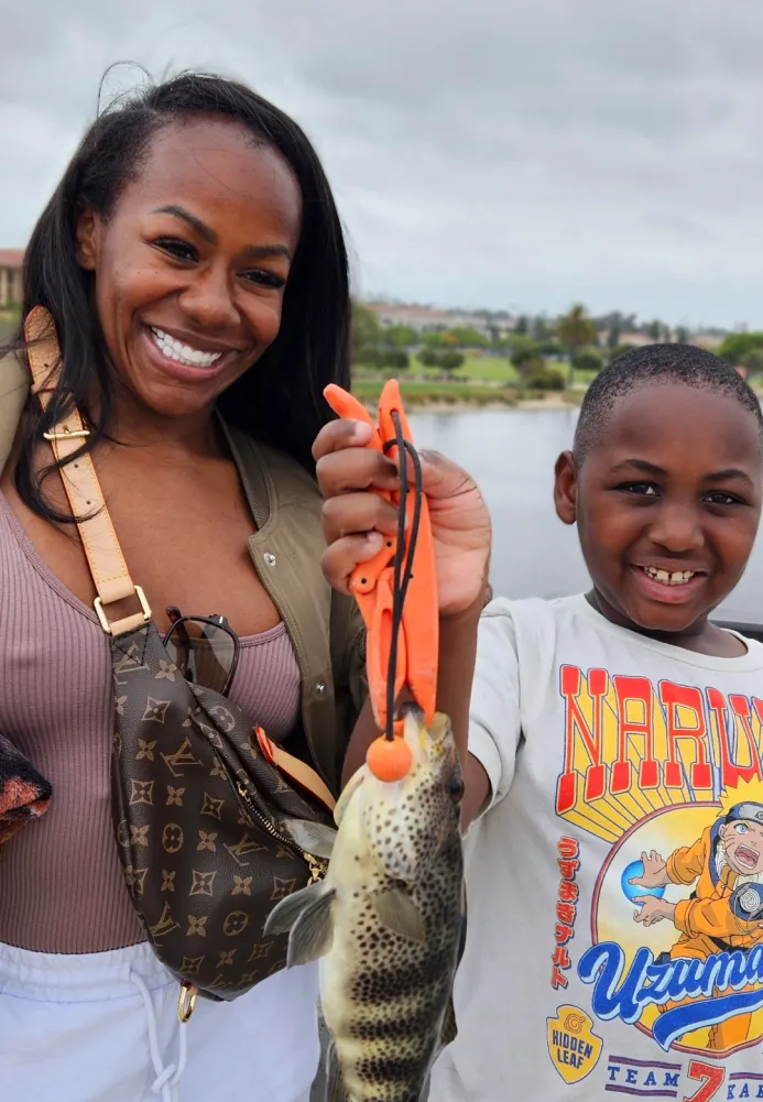 fishing-lessons-1 Smiling woman and boy holding a fish with pliers by a lake, cloudy sky in the background.