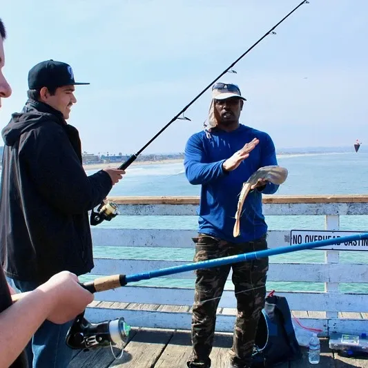 Three people fishing on a pier, one holding a fish. Ocean and coastline visible in the background.