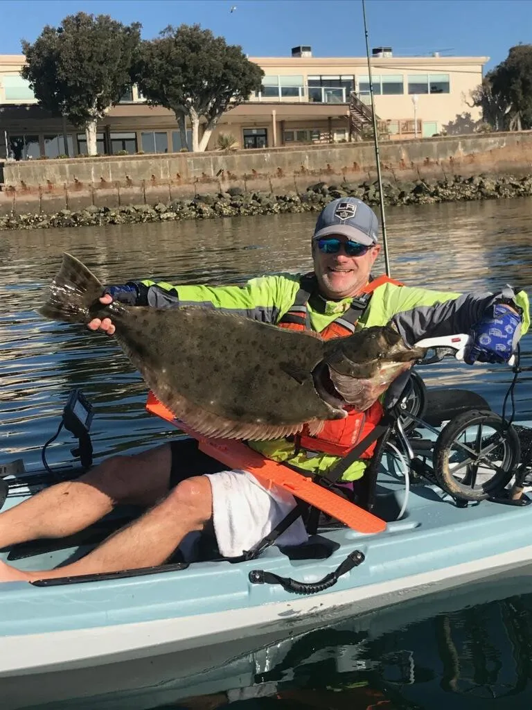kayak-fishing-2 (2) Person on a kayak holding a large fish, with buildings and trees in the background.
