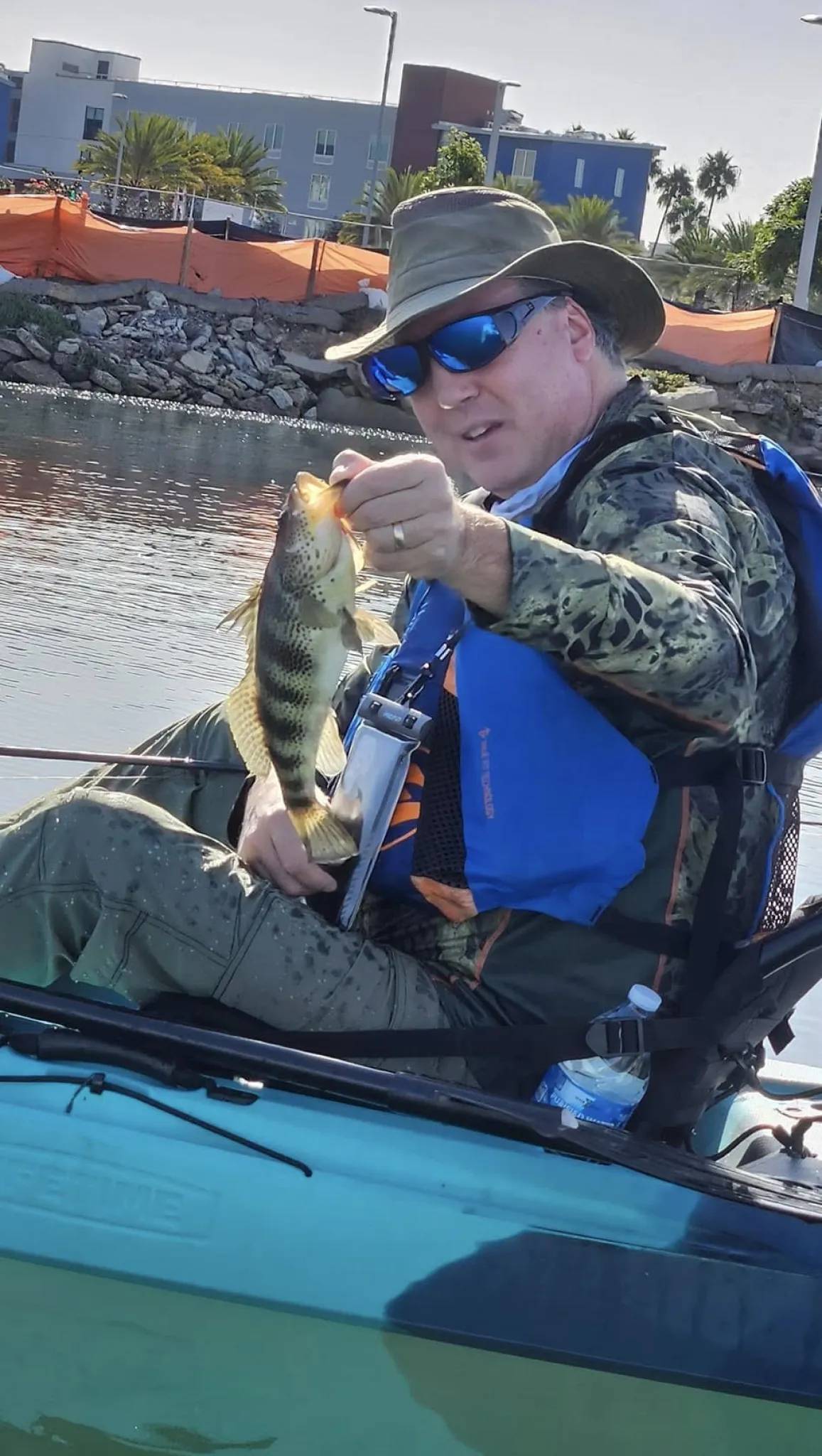 Person in a kayak holds up a fish, wearing a camo outfit, blue life vest, and sunglasses on a sunny day.
