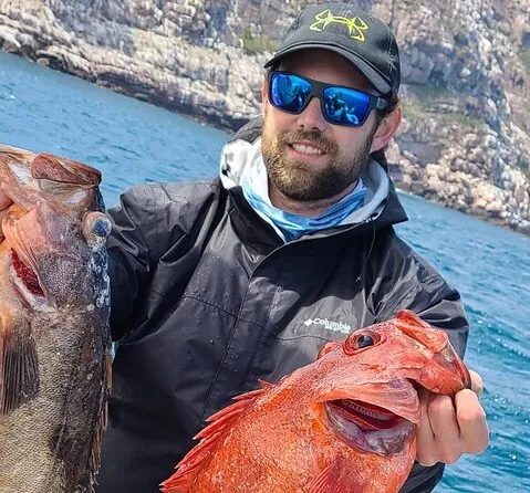 Man in sunglasses and cap holds two fish on a boat with rocky cliffs in the background.