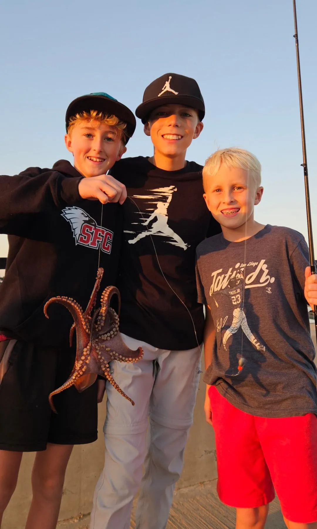 9EC4087E-B56D-4CD2-B485-F66129F76868 Fishing Camp in San Diego: Three boys smile at sunset; one holds a small octopus, another holds a fishing rod.
