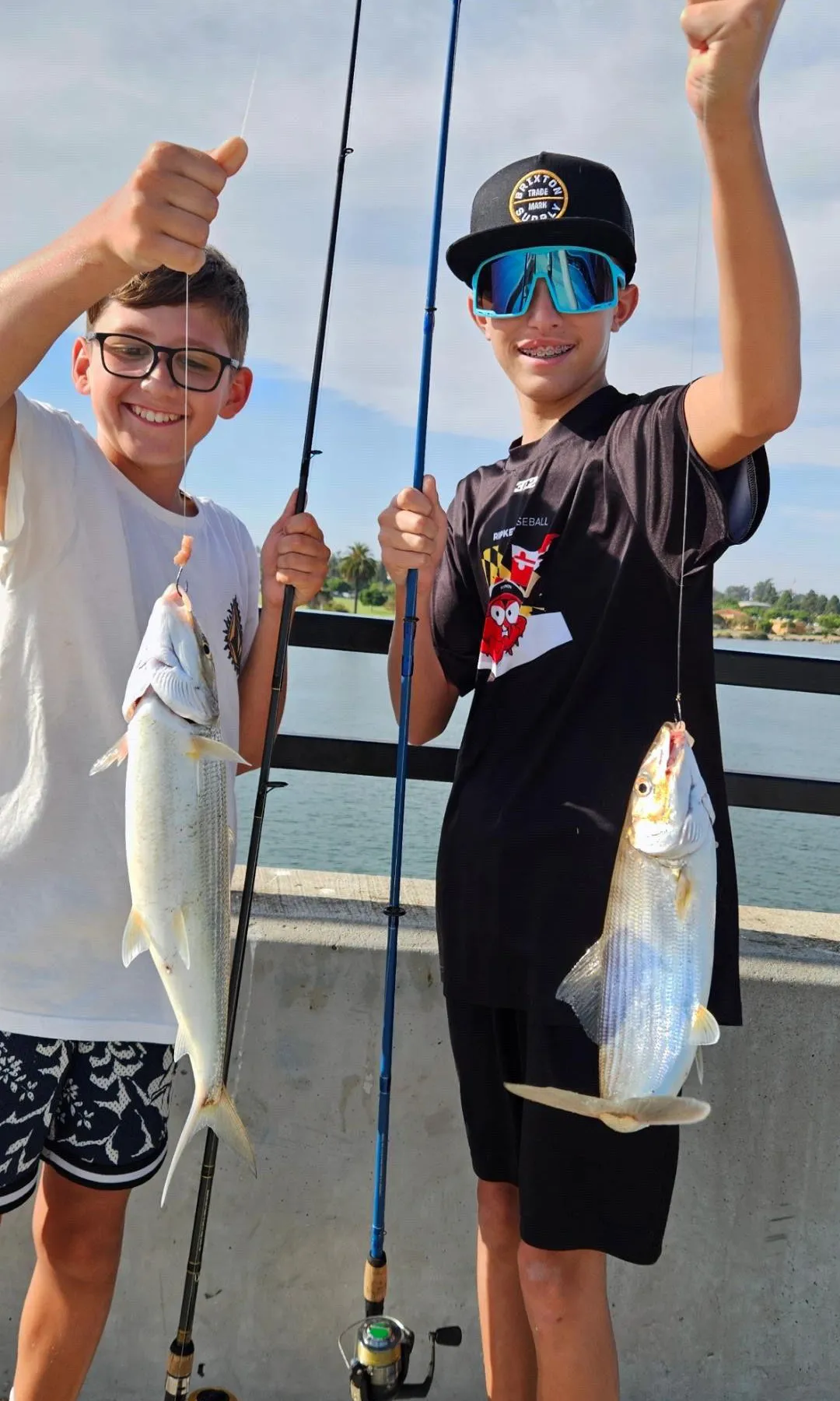 D5B3652F-563D-457C-AF27-F50E0B31D593 Two boys smiling and holding fishing rods with freshly caught fish by the water on a sunny day.