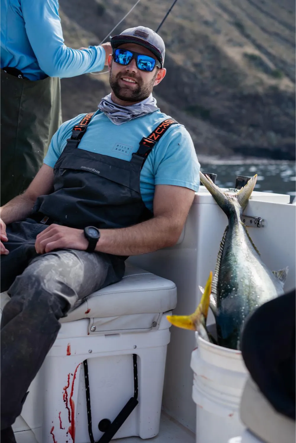 deep-sea-fishing-1 Man in fishing gear sits on a cooler on a boat, with a large fish in a bucket and blood on the deck.