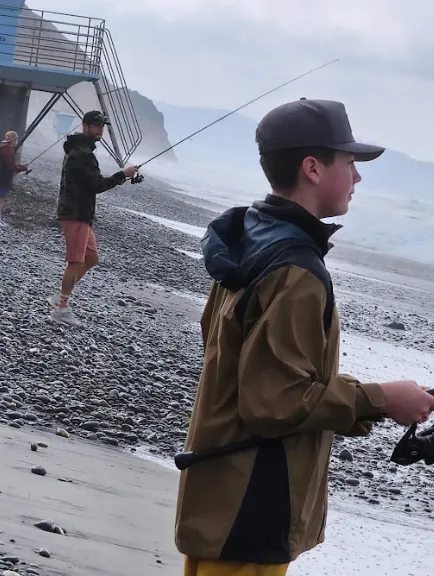 fishing-lessons-1 Two people wearing jackets and hats fish on a rocky beach by the ocean, with waves in the background.