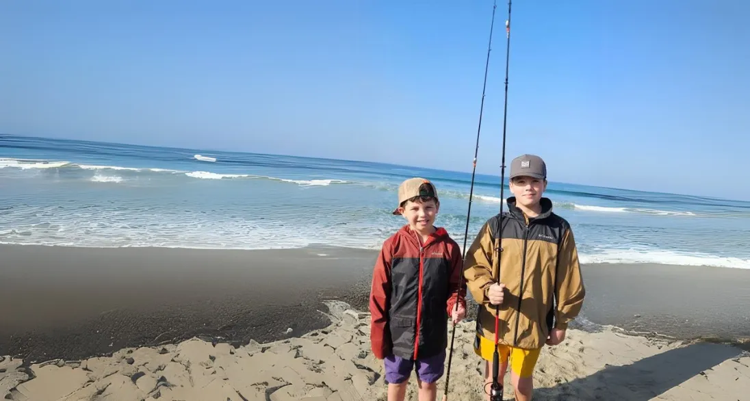 Two boys holding fishing rods stand on a sandy beach with waves and blue sky in the background.