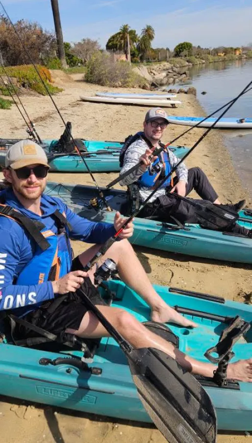 Two men sit in blue kayaks on a sandy shore, holding fishing rods and smiling at the camera.