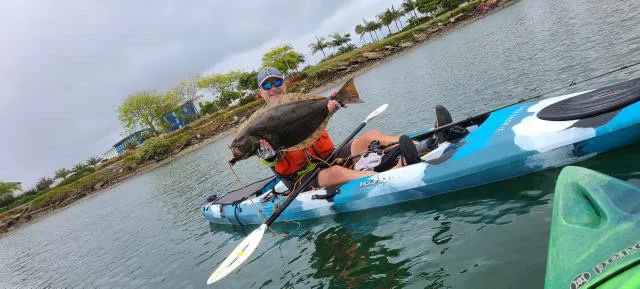 kayak-fishing-h Person in a blue kayak holds up a large fish while on calm water near a tree-lined shore.