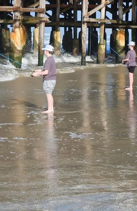 shoreline-fishing-1 Two people fish while standing barefoot in wet sand near a wooden pier with waves approaching.