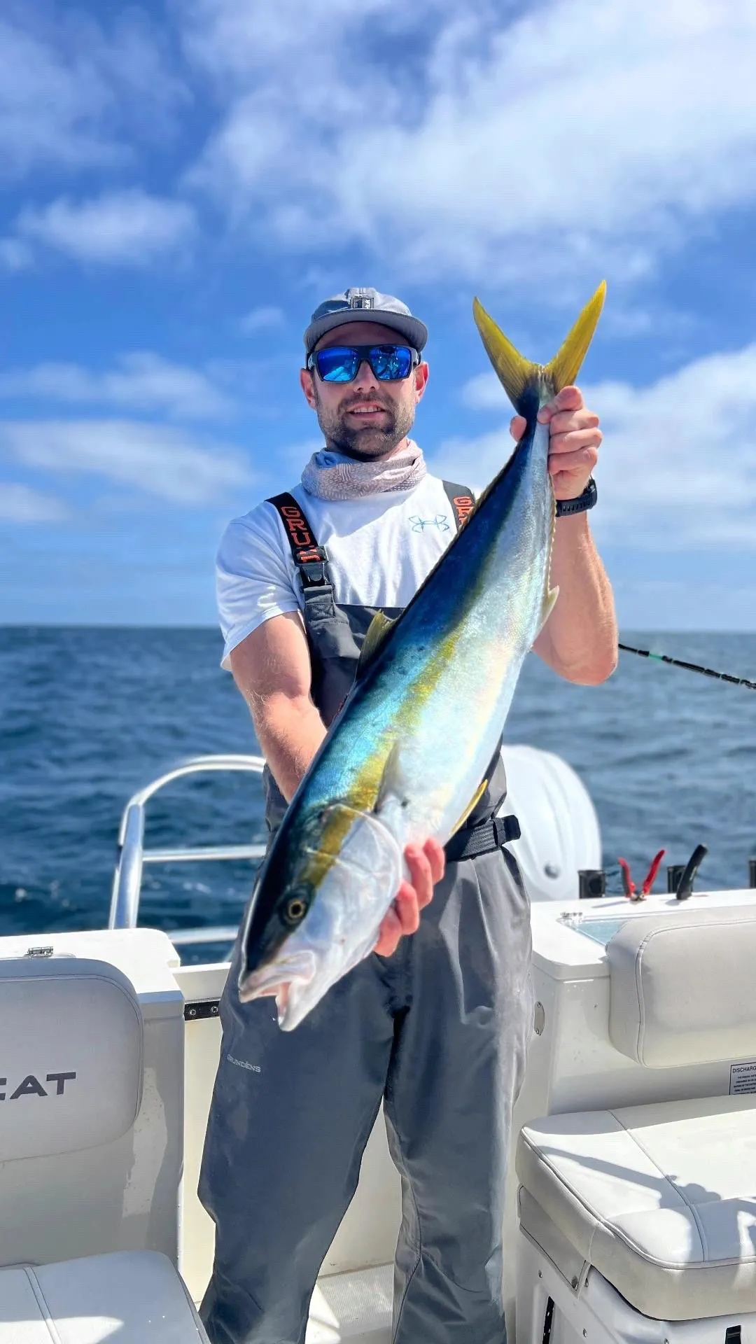 sports-fishing-1 Man on a boat holding a large yellowtail fish with the ocean and blue sky in the background.