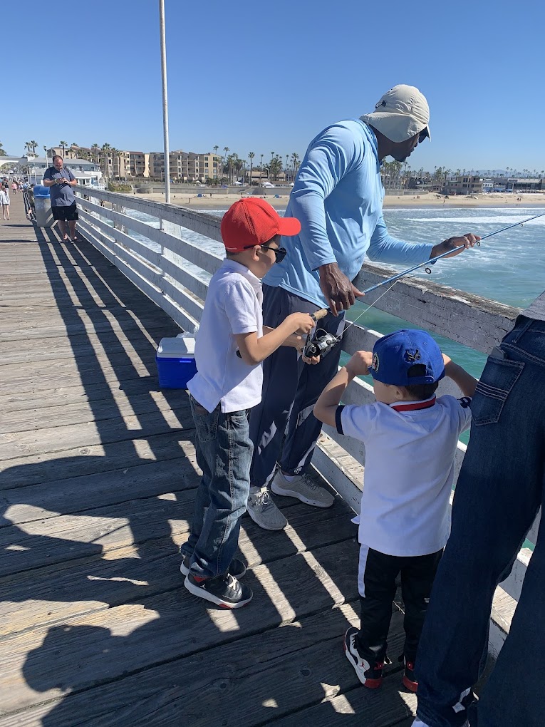 unnamed Two young boys fishing with an adult on a sunny pier, looking out over the ocean.