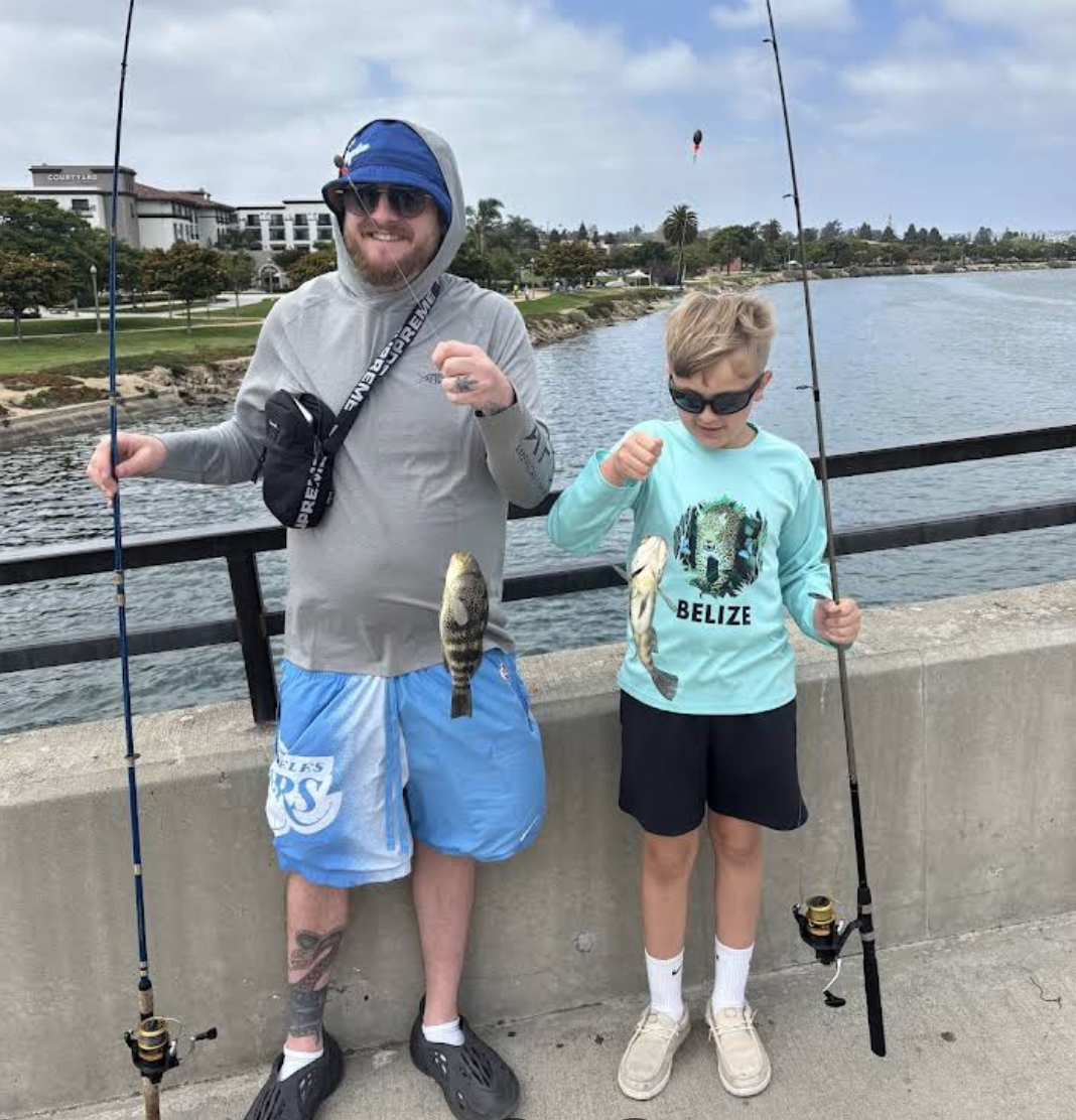 Captura de pantalla 2025-10-08 a la(s) 12.47.35 p.m. Man and boy smiling on a bridge, each holding a fishing rod and a freshly caught fish.