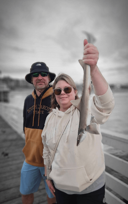 Woman in sunglasses holds up a fish on a pier, with a man standing beside her. Background is in black and white.