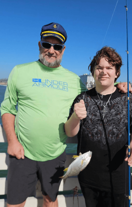 An older man and a teen smiling on a boat; the teen holds a fishing rod with a small fish.