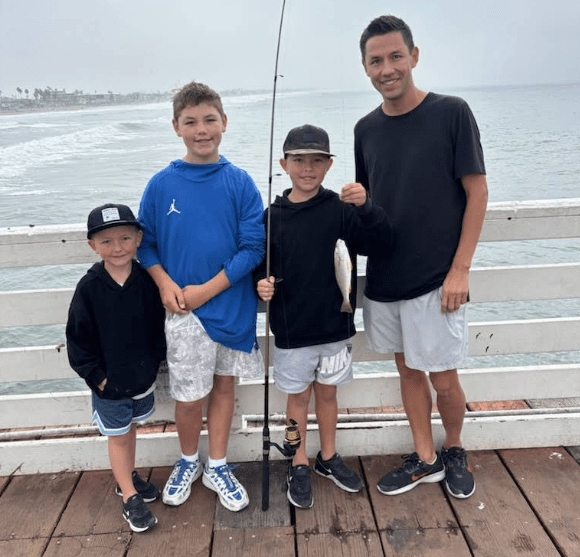 Four people stand on a pier by the ocean, one boy holding a fishing rod with a small fish.