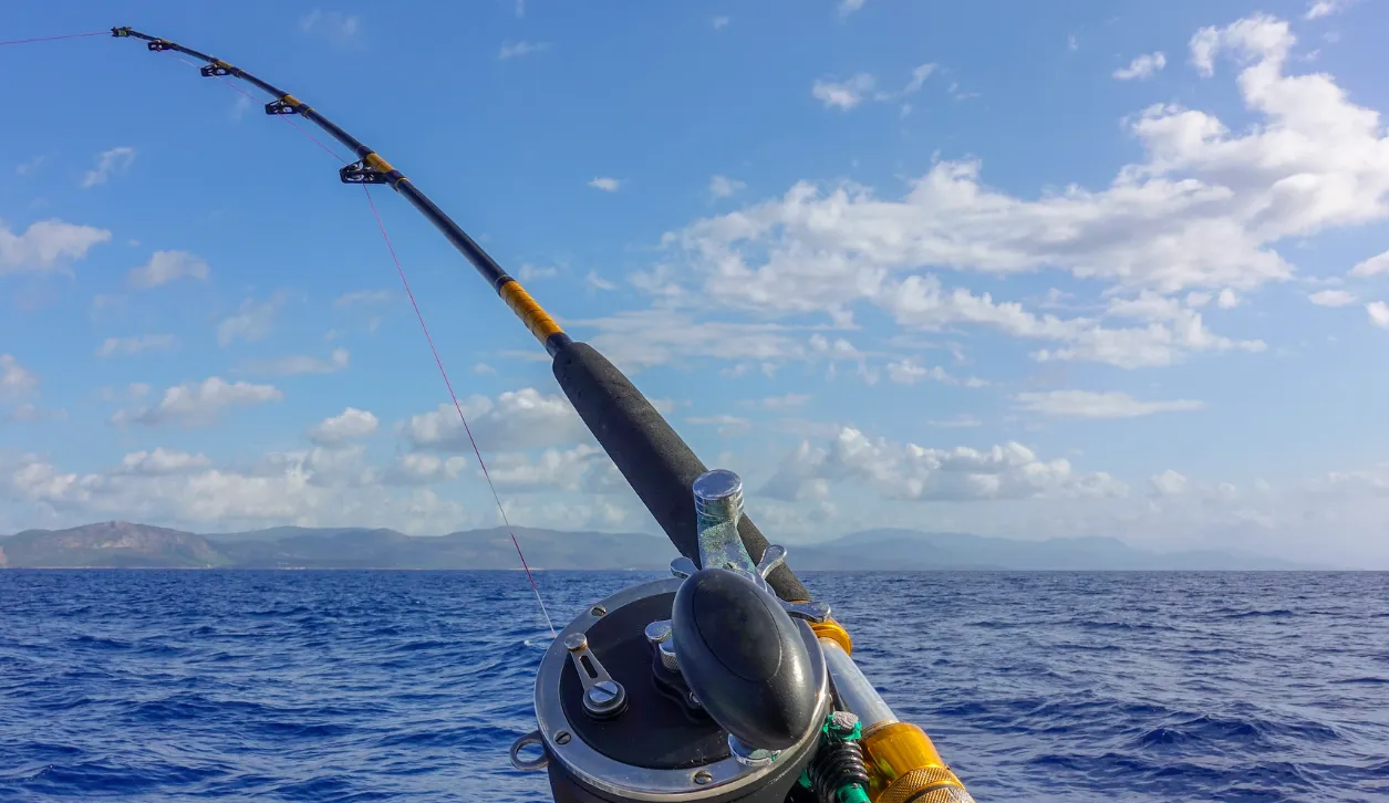 Close-up of a fishing rod over deep blue ocean water with distant mountains and a partly cloudy sky.