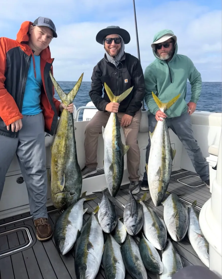 Three people on a boat hold large fish, with more fish laid out on the deck, smiling at the camera.