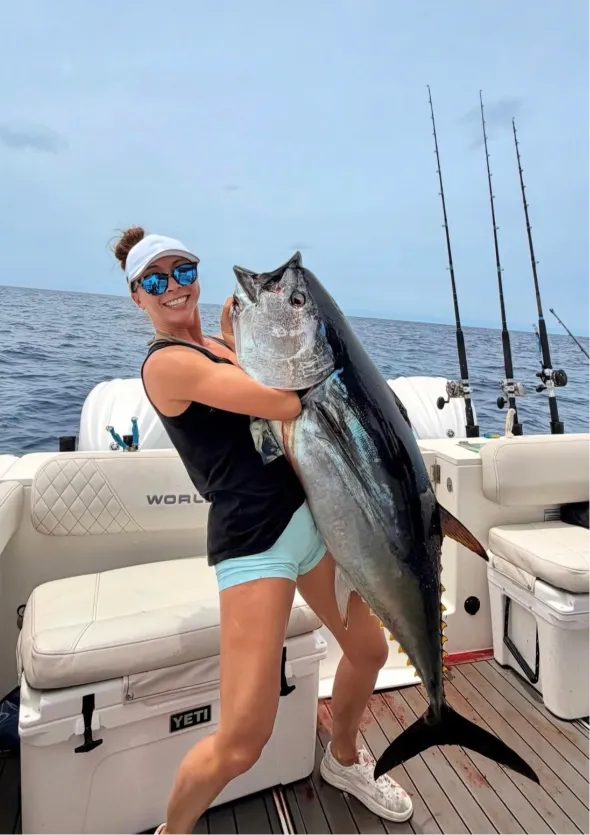 Woman on a boat holding a large fish, smiling, with fishing rods and ocean in the background.